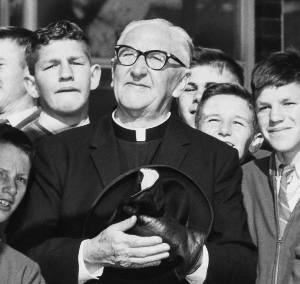 A closeup of Father Dunlea surrounded by a group of smiling boys at Boys Town in Engadine, now known as the Dunlea Centre. Father Dunlea holds his hat and gloves in his hands in a moment of reflection. 